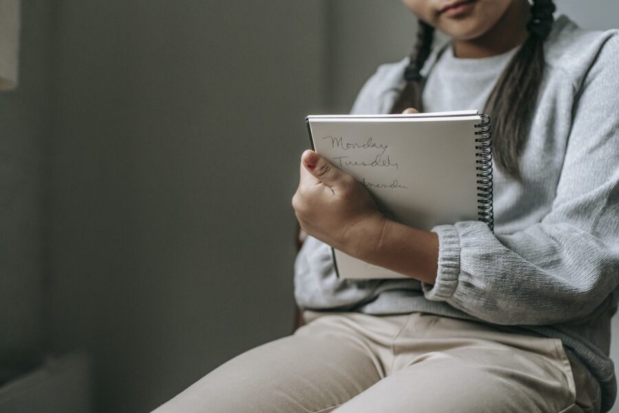 Crop unrecognizable girl in casual outfit sitting on chair and taking notes in copybook