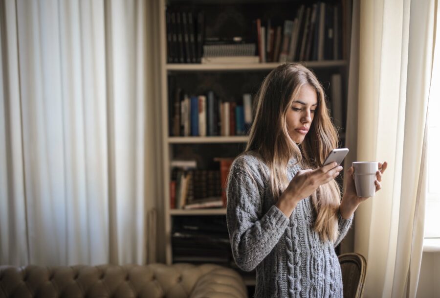 Photo of Woman in Gray Sweater Holding White Ceramic Cup While Using Her Phone